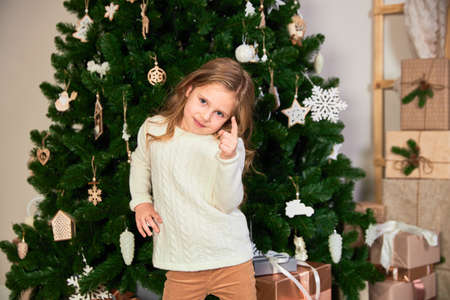 A girl in white eco dress dressed up as a Christmas girl. Theres a green Christmas tree and presents in the back. Girl points in camera.の写真素材