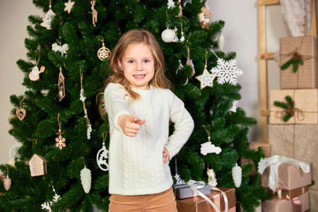 A girl in white eco-clothes stands against the background of a green Christmas tree and gifts. The girl points to the camera.の写真素材