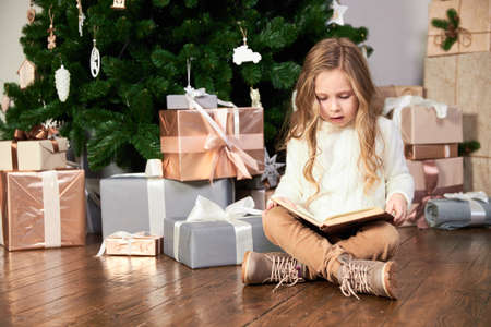 A girl in white eco clothes sits next to a Christmas tree, presents and reads a bookの写真素材