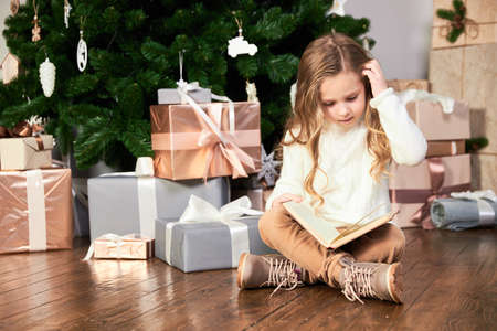 A girl in white eco-clothes sits next to a Christmas tree and reads a book, scratches her head and thinksの写真素材