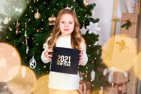 A little cute girl holds a black gift with a gold inscription 2021, at the back of a Christmas treeの写真素材