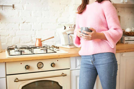 A woman in a light kitchen loft near a vintage slab stands with a cup in her hands, no face, only...
