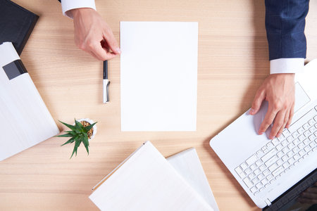 A wooden office desk with a laptop and a white sheet for recording. View from above at the hands of a businessman with a pen, ready to write. Flatley with business accessories.の写真素材