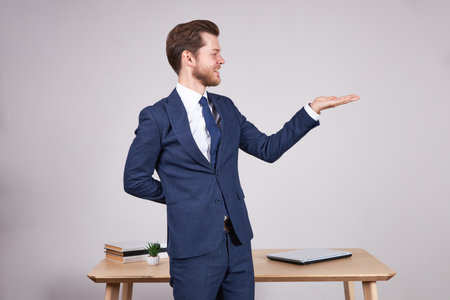 A businessman in a blue business suit looks near a wooden office table with an open palm. White background.の写真素材