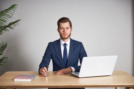 A man businessman in a blue suit with a laptop at his desk looks into the camera with a serious faceの写真素材
