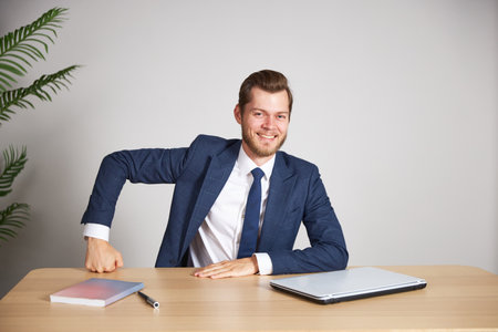 A businessman in a blue business suit sits at his desk clutching his hand in his fist. He looks directly into the camera and smiles. White background.の写真素材