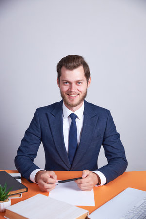 The businessman sits at an orange office desk, holding a pen. On the table is a laptop, books and a white sheet of paper. The man looks at the camera and smiles. White background.の写真素材