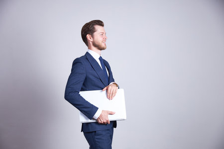 A man in a blue office suit holds a white laptop, looking to the side. White background.の写真素材