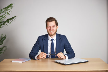 A man businessman in a blue business suit sits at his desk, smiling and holding a pen. White background.の写真素材