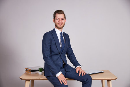 A businessman in a blue business suit sits on an office wooden table, looks into the camera and smiles. White background.の写真素材
