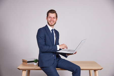 A businessman in a blue business suit sits on a wooden office table and holds a laptop, look at the camera and smile. White background.の写真素材