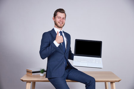 A businessman in a blue business suit sits on an office desktop, holds a laptop with a black screen and shows his thumb up.の写真素材