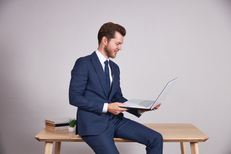 A businessman in a blue business suit sits on a wooden office table and holds a laptop, look at the screen. White background.の写真素材