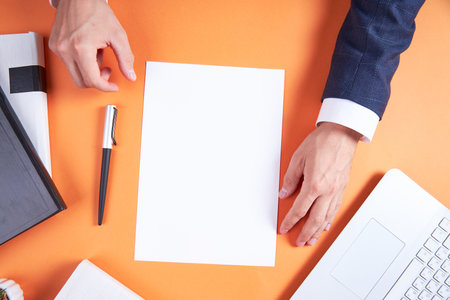Orange office desktop with business accessories. Laptop, books, pen. The businessman is holding a white sheet of paper with an empty background. Flatley is a business of details.の写真素材