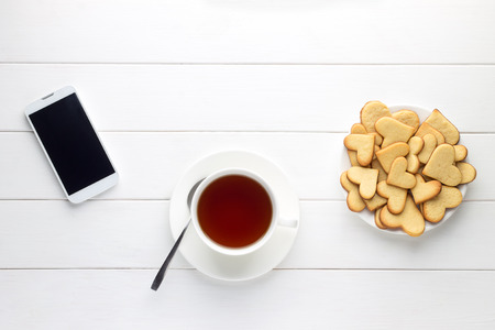 Breakfast of cup of tea and heart shaped cookies and smartphone on wooden table. Morning news reading on the phone. Top view, copy space.の写真素材
