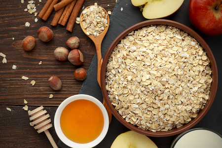 Ingredients for healthy breakfast: rolled oat flakes, milk, honey, hazelnut, cinnamon, apple on dark rustic wooden table. Top view.の写真素材