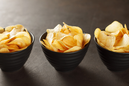 Three black bowls of potato chips on brown table. Tasty junk crunchy food with copy space.の写真素材