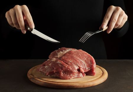 Hands holding fork and knife and ready to eat raw pork meat on the wooden cutting board on dark background. Nonvegetarian concept, copy space.の写真素材