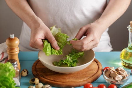 Chef tears the lettuce leaves into ceramic bowl on kitchen table with fresh ingredients.の写真素材