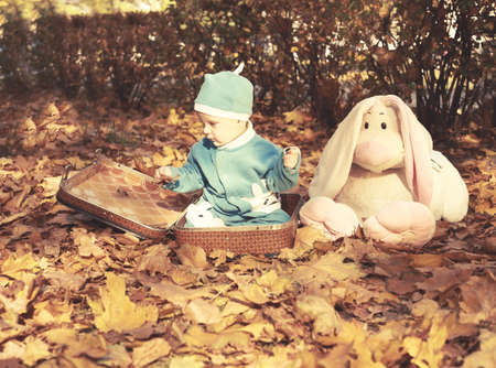 Little boy in nature sitting in open suitcase. Outings - the forest in autumn. Nearby are big hare toyの写真素材