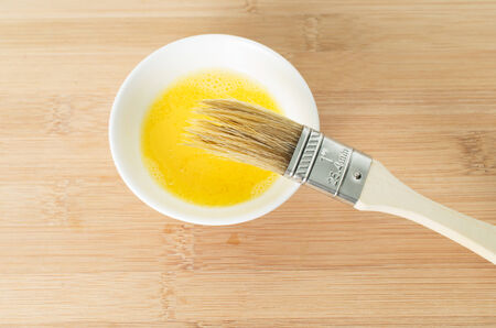 Wooden brush dipped into the whisked egg white and egg yolk in a small ceramic bowl during preparation of pastry の写真素材