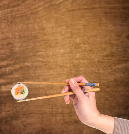 Woman hand holding fresh maki sushi roll with wooden chopsticks against wooden background with copy...