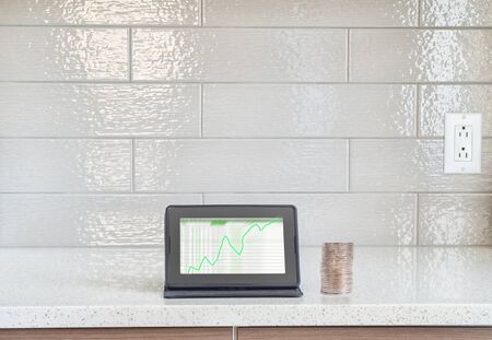 Digital tablet with green stocks screen and stack of coins on a granite counter top