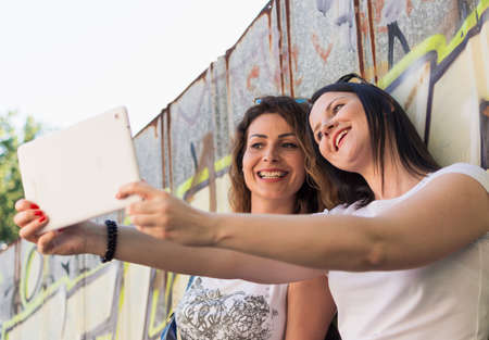 two adult  girls sitting in the shade on the stairs, listening to music and photographing with a tabletの写真素材