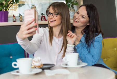 portrait of two adult smiling girls taking photographs with a mobile phone indoorの写真素材