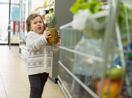 a little cute little girl holds the pineapple in her hands and wants to put it in a shopping cartの写真素材