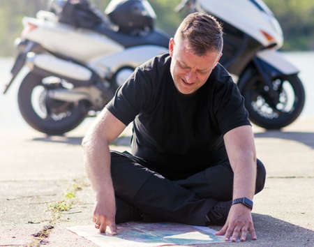 a mature man takes a brake sitting in front of a motor scooter and studies a driving map of the area...