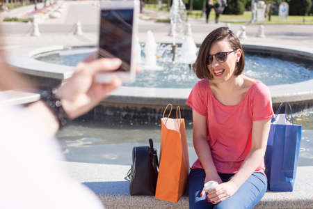two adult girls sitting in front of a water fountain and photographing with a mobile phone.Spending a sunny summer day in the cityの写真素材