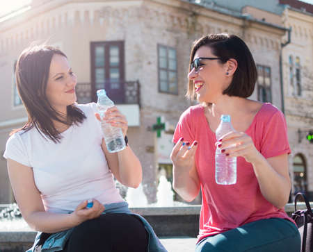 two adult girls drink water from a bottle while sitting in front of a water fountain on a warm, sunny dayの写真素材