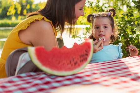 A daughter and mother sit at the table and eating watermelon slice. Girls sit at the table and eating watermelon slice. A mother cleaning daughter with the napkin after sweet fruit. Fruit consuming.の写真素材
