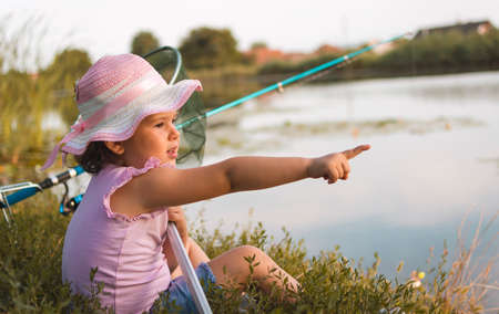 Sweet little girl sitting on the grass and catching the fish near  the river. Little girl, 4-5 years, spending fishing time alone on the river coast. Kids and fishing.の写真素材