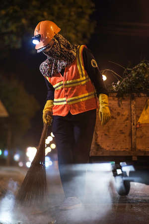 A woman in a protective suit with a broom cleans the street at night.の写真素材