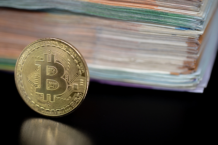 Bitcoin token on black with reflection, with a big pile of euro banknote money in the background. Cryptocurrency versus paper currency conceptの写真素材