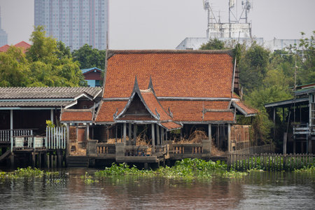 A traditional Thai wooden house on stilts by a river, featuring a distinctive red-tiled roof and surrounded by lush greenery and water lilies. The scene captures the serene beauty of Thai architecture and nature.の写真素材