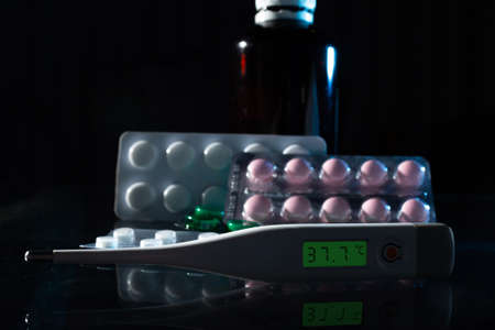 medical jars, thermometer, syringe, gauze mask and pills medicines, medicine on a thermometer 37.7 degrees Celsius on glass black background close up. copy spaceの写真素材