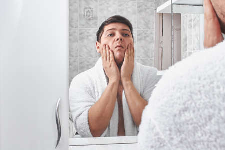 a man in a white bathrobe looks in the mirror in the bathroom.の写真素材