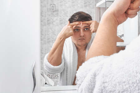 a man in a white coat squeezes acne on his face through a mirror in the bathroom.の写真素材