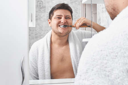 a man in a white bathrobe brushing his teeth through a bathroom mirror.の写真素材