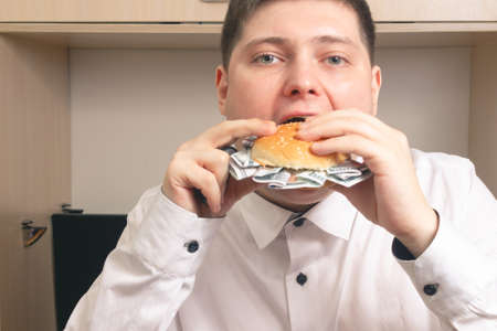 Caucasian man eating a burger with hundred dollar bills in a white shirt in the office