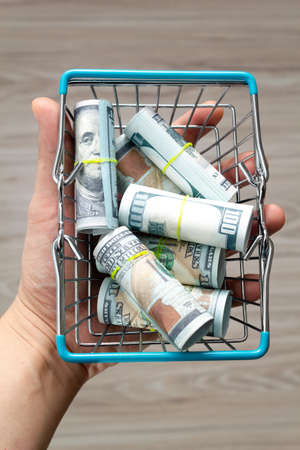 shopping basket in hand with hundred dollar rolls on a light parquet backgroundの写真素材