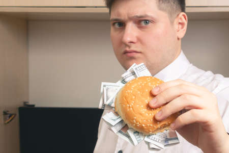Caucasian man holds a burger with hundred dollar bills in a white shirt in the officeの写真素材