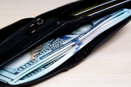 a bundle of hundred-dollar banknote in a black leather wallet on a light wooden. view from the side. close upの写真素材