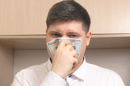 Caucasian man blows his nose wipes his nose with a hundred dollar bill in a white shirt in the officeの写真素材