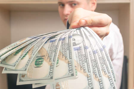 Caucasian man holds out to the camera a bundle of hundred-dollar bills in a white shirt in the officeの写真素材