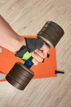 a male hand is on a glove with a dumbbell against a background of a quarantine fitness mat at home on a light wooden floorの写真素材