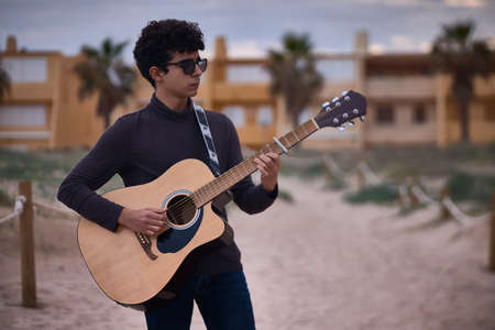 young caucasian teenage boy playing acoustic guitar on the beach. sunglasses and dark clothing.の写真素材